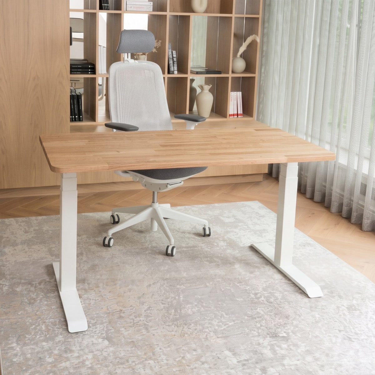 A modern adjustable height standing desk with a light oak wood top and white metal legs, styled in a home office with a white mesh office chair, wooden shelving, and a patterned rug.