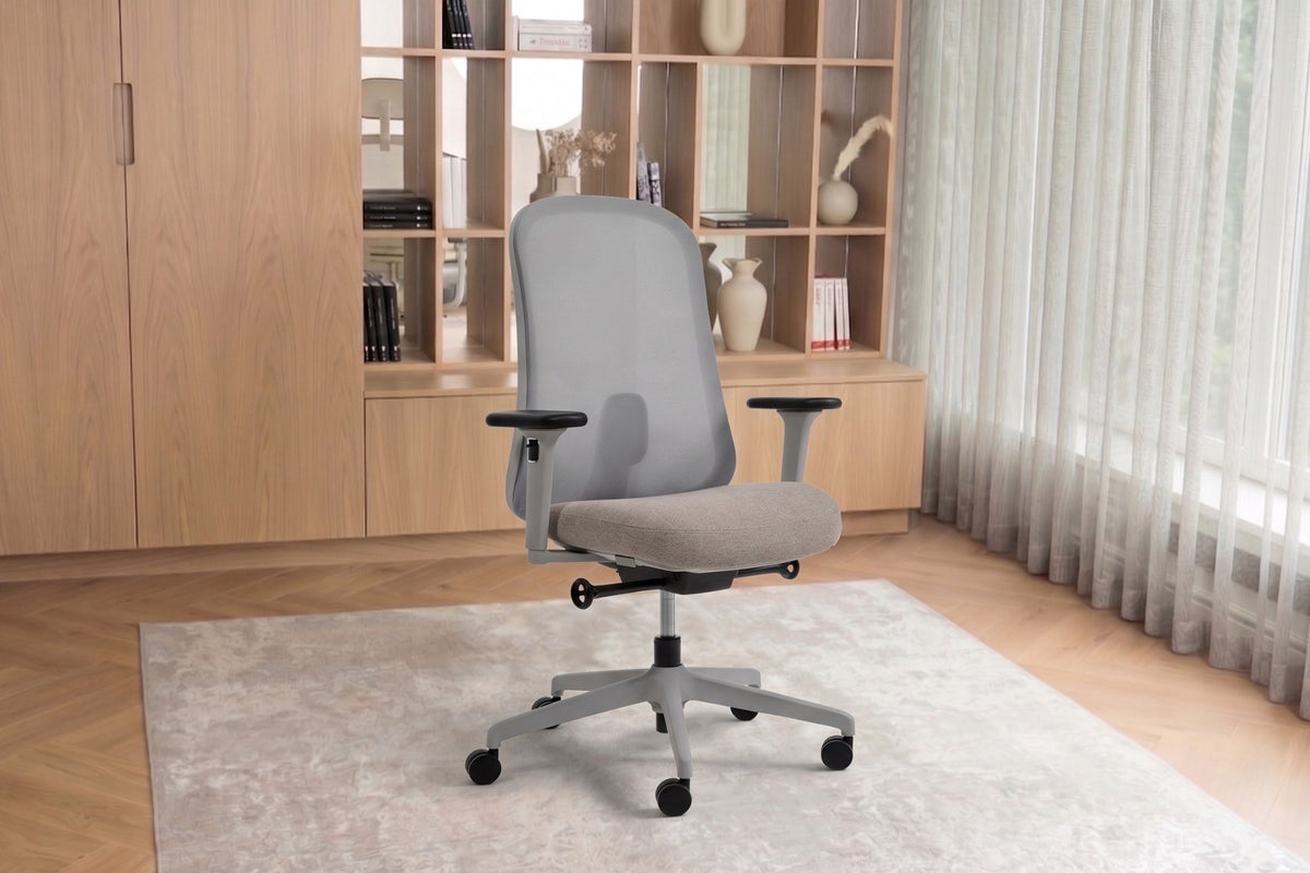 A modern light taupe fabric office chair with a five-star wheeled base, positioned on a light rug in a bright, contemporary home office setting with wooden shelving and sheer curtains.