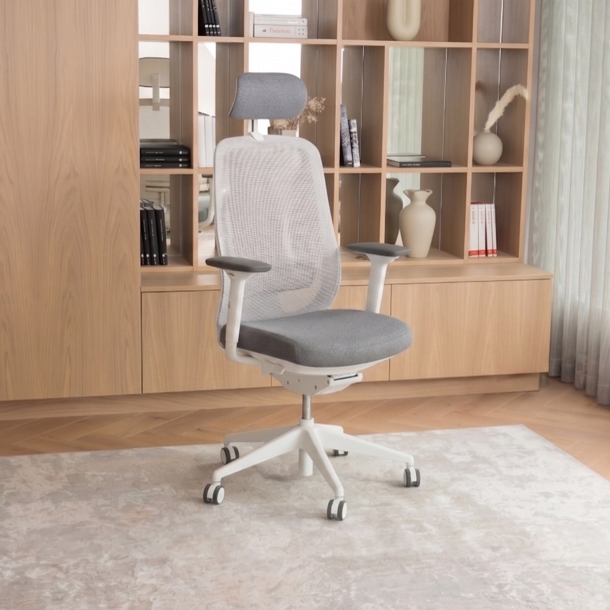 A modern gray mesh office chair with a white base and five-star wheel legs, positioned on a light-colored rug in front of a wooden bookshelf.
