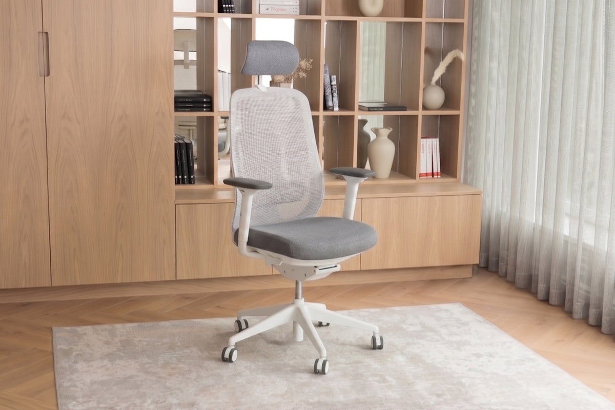 A modern ergnomic light gray mesh office chair with a white five-star base and casters, positioned on a light rug in front of a wooden bookshelf and cabinet in a bright room.