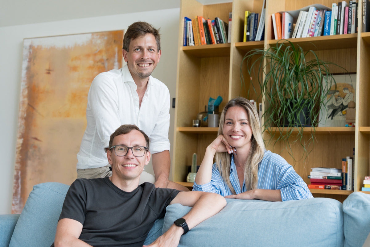 Three smiling people in a bright, modern living space with a light blue sofa, wooden bookshelves, and a large abstract painting on the wall.