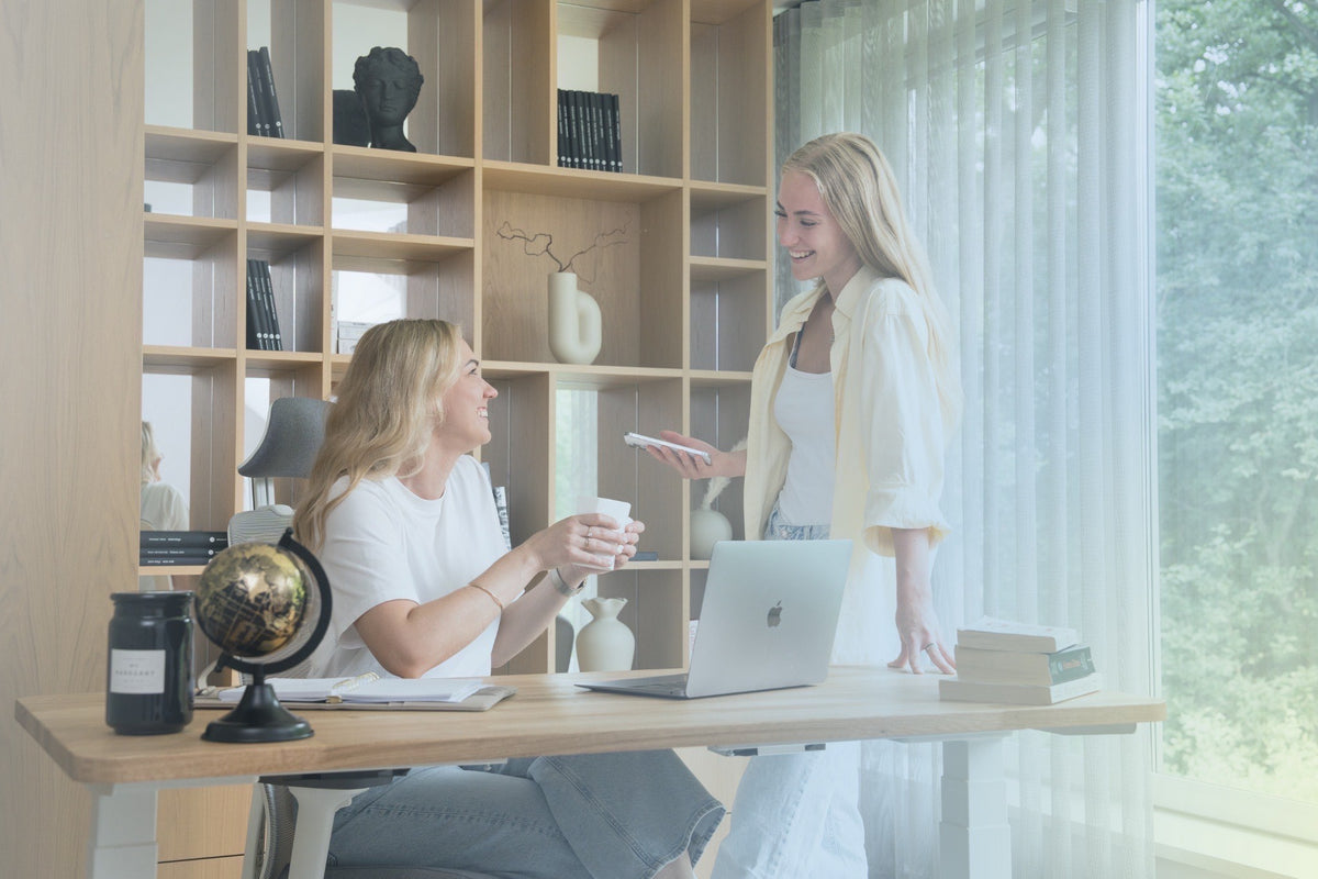 Two women working in a bright, modern home office. One woman is seated at a wooden height-adjustable desk with a laptop and coffee cup, while the other stands beside her holding a phone.
