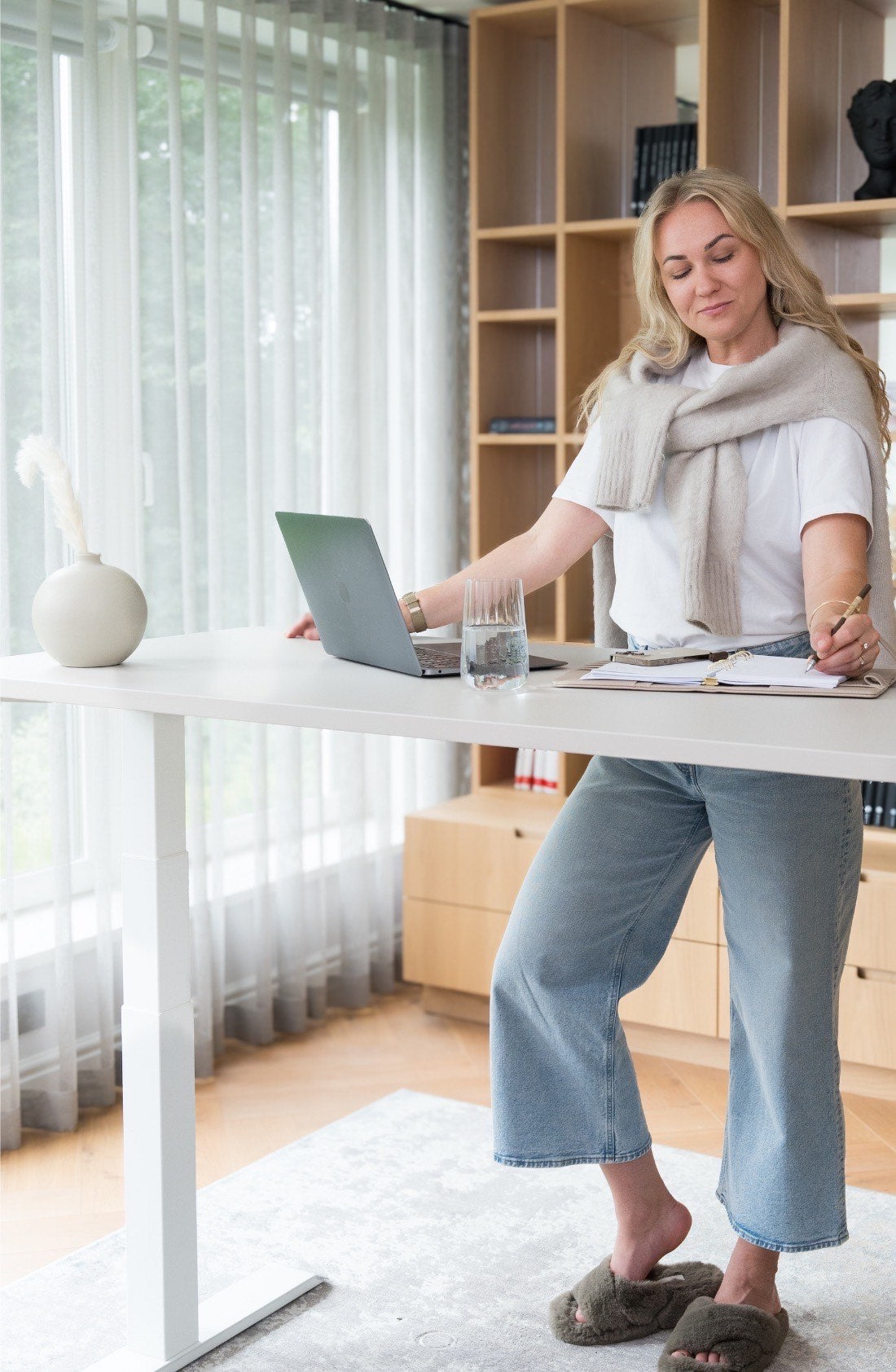 Woman stands at a white height-adjustable standing desk in a bright home office, using a laptop and writing in a notebook. 